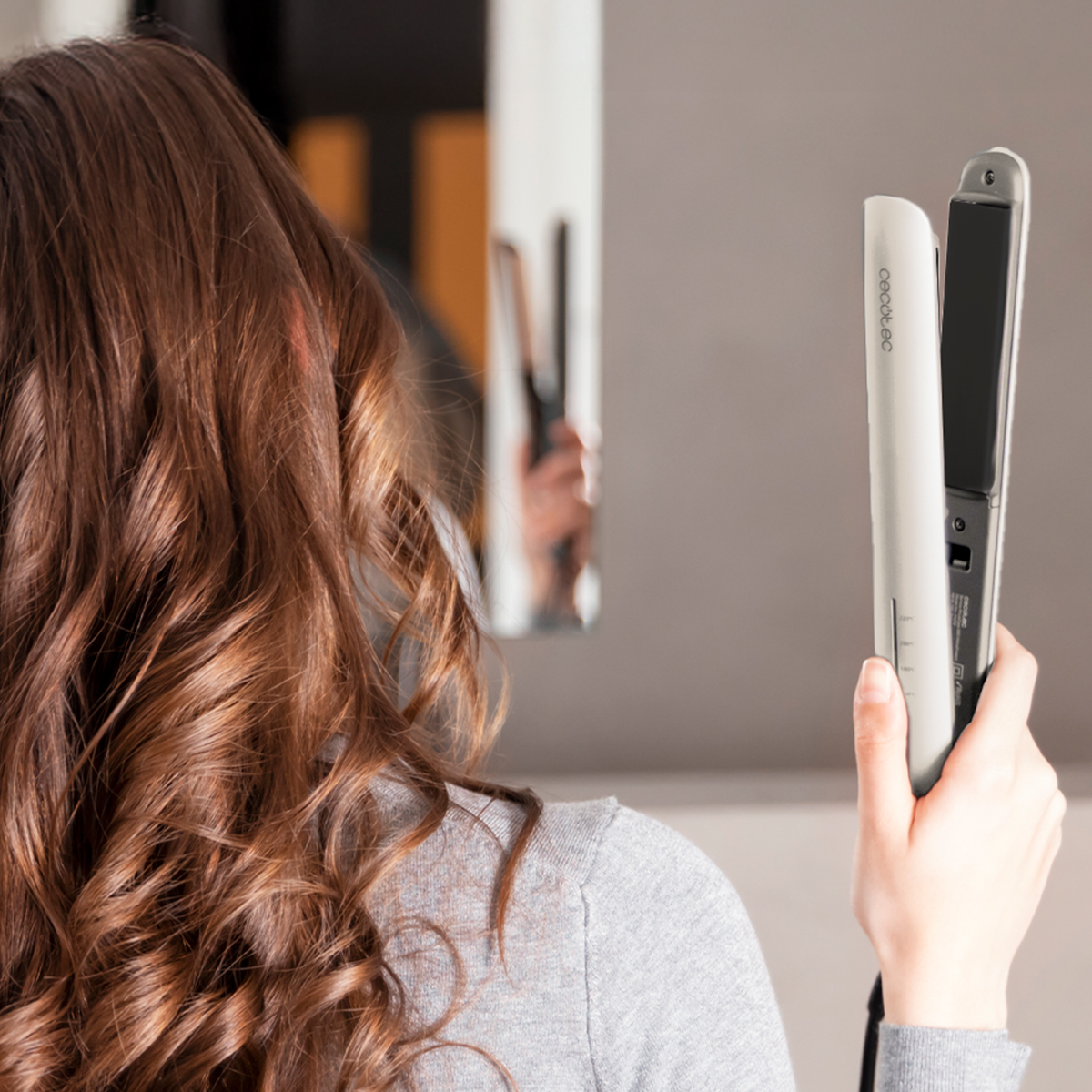 Una mujer sostiene una plancha para el cabello blanca. Se ven su cabello castaño rizado y una camiseta gris. Un espejo refleja la plancha.