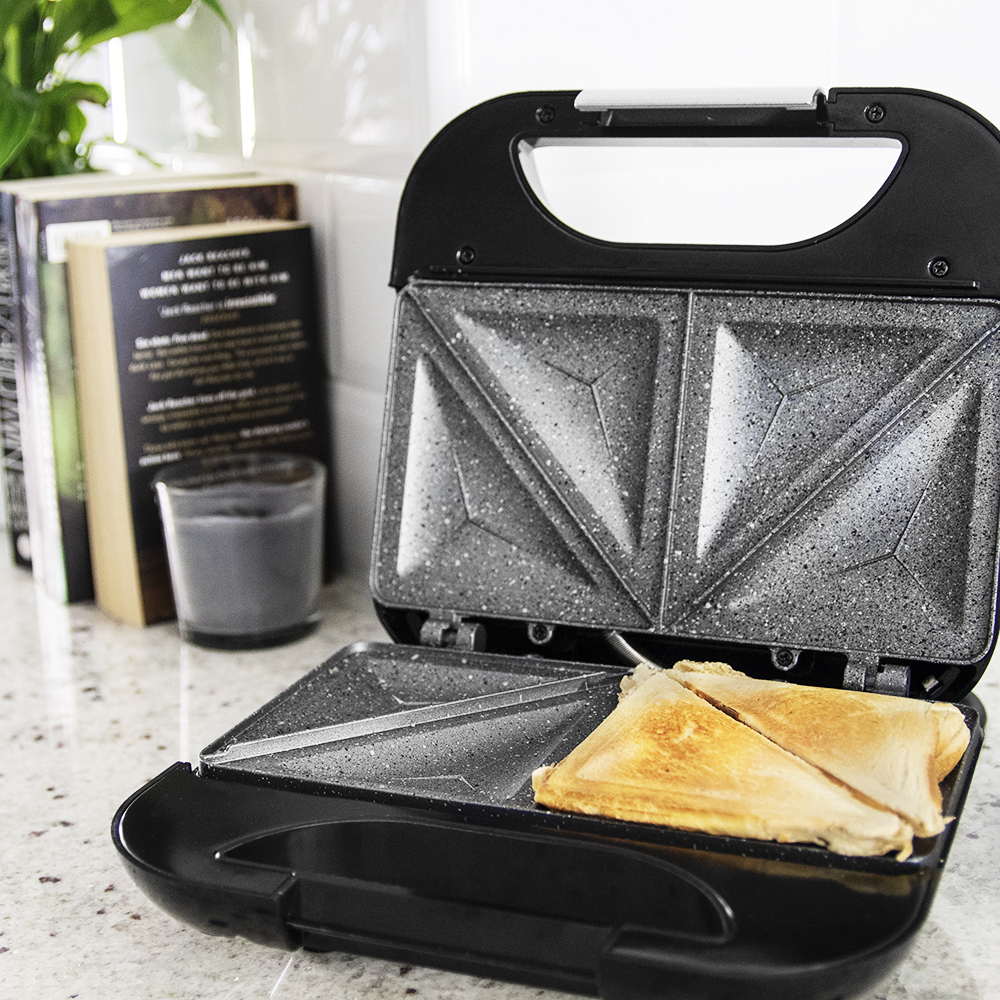 Sandwichera abierta con dos mitades de sándwich cocinadas. Libros y una vela en el fondo.
