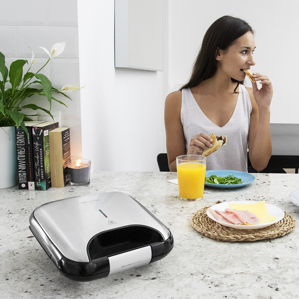 Mujer comiendo un sándwich. Una sandwichera, zumo e ingredientes en la mesa. Plantas y libros al fondo.