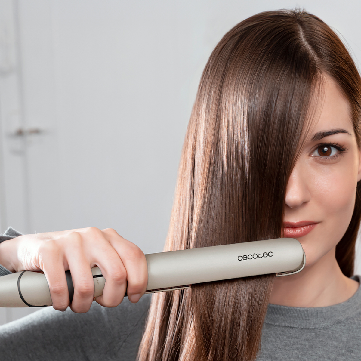 Una mujer alisa su cabello castaño con una plancha plateada. Lleva una camisa gris. El fondo es una pared blanca.