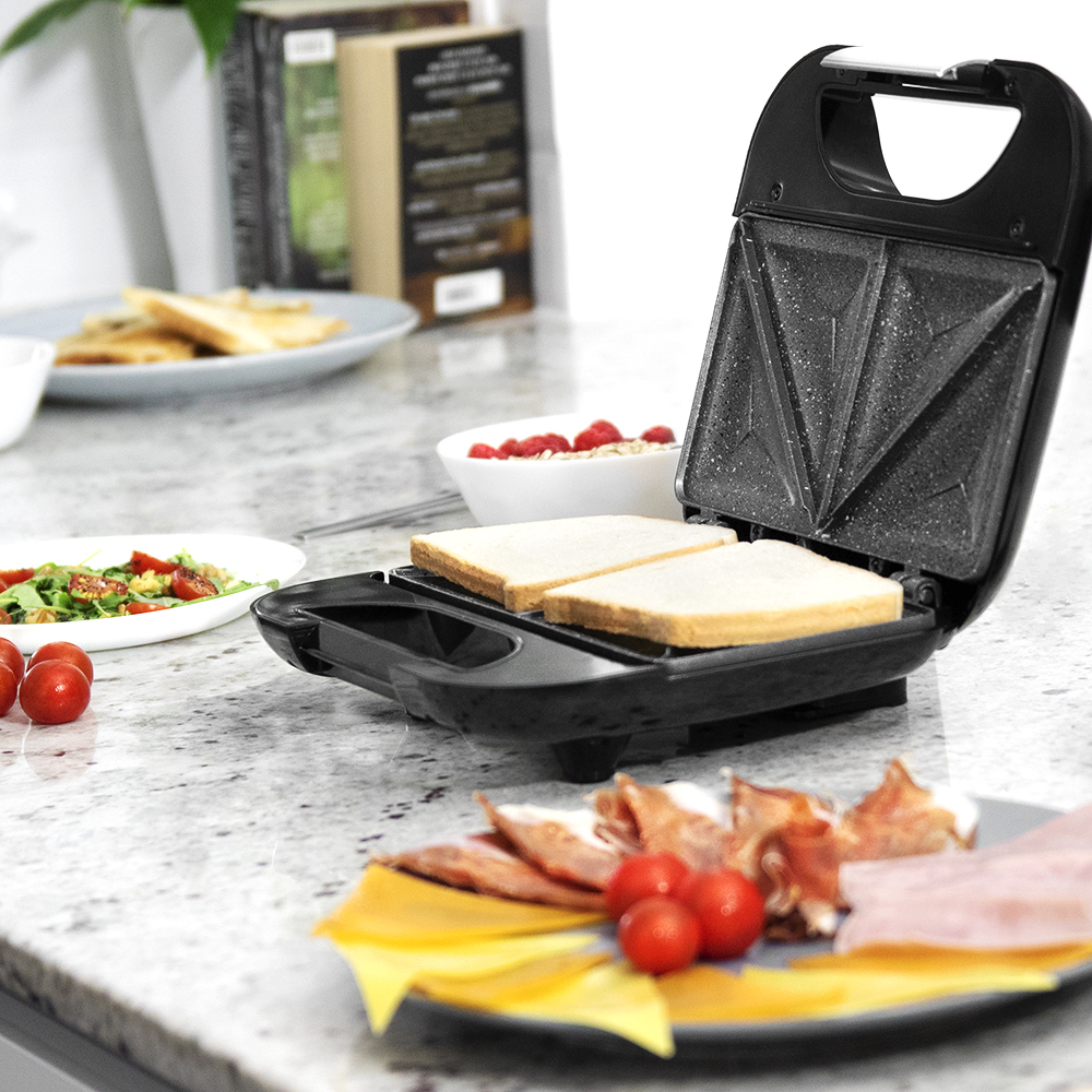 Una sandwichera con pan dentro. Un plato tiene queso, jamón y tomates. Otros platos tienen ensalada y sándwiches con frambuesas.