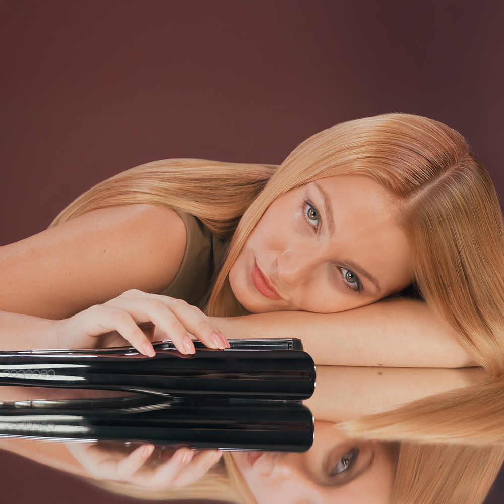 Una mujer rubia se apoya en una superficie de espejo. Una plancha negra para el cabello está al frente. El fondo es marrón.