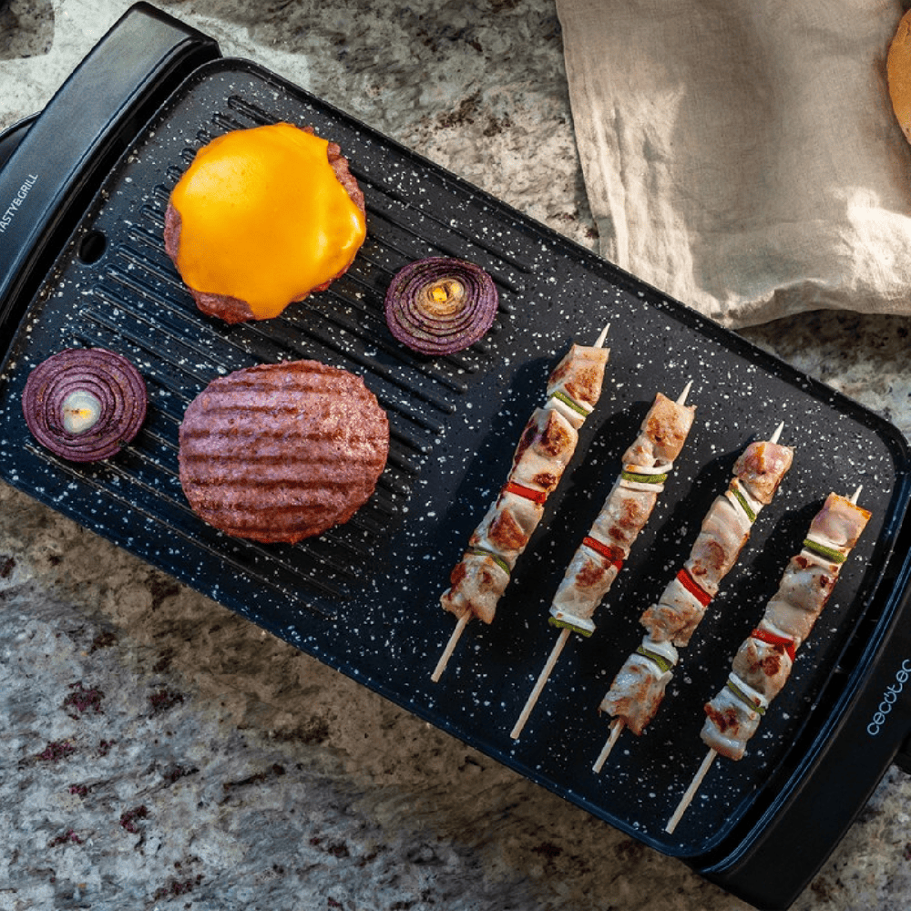Una parrilla negra con hamburguesa, queso, cebollas rojas y brochetas de carne y verduras.