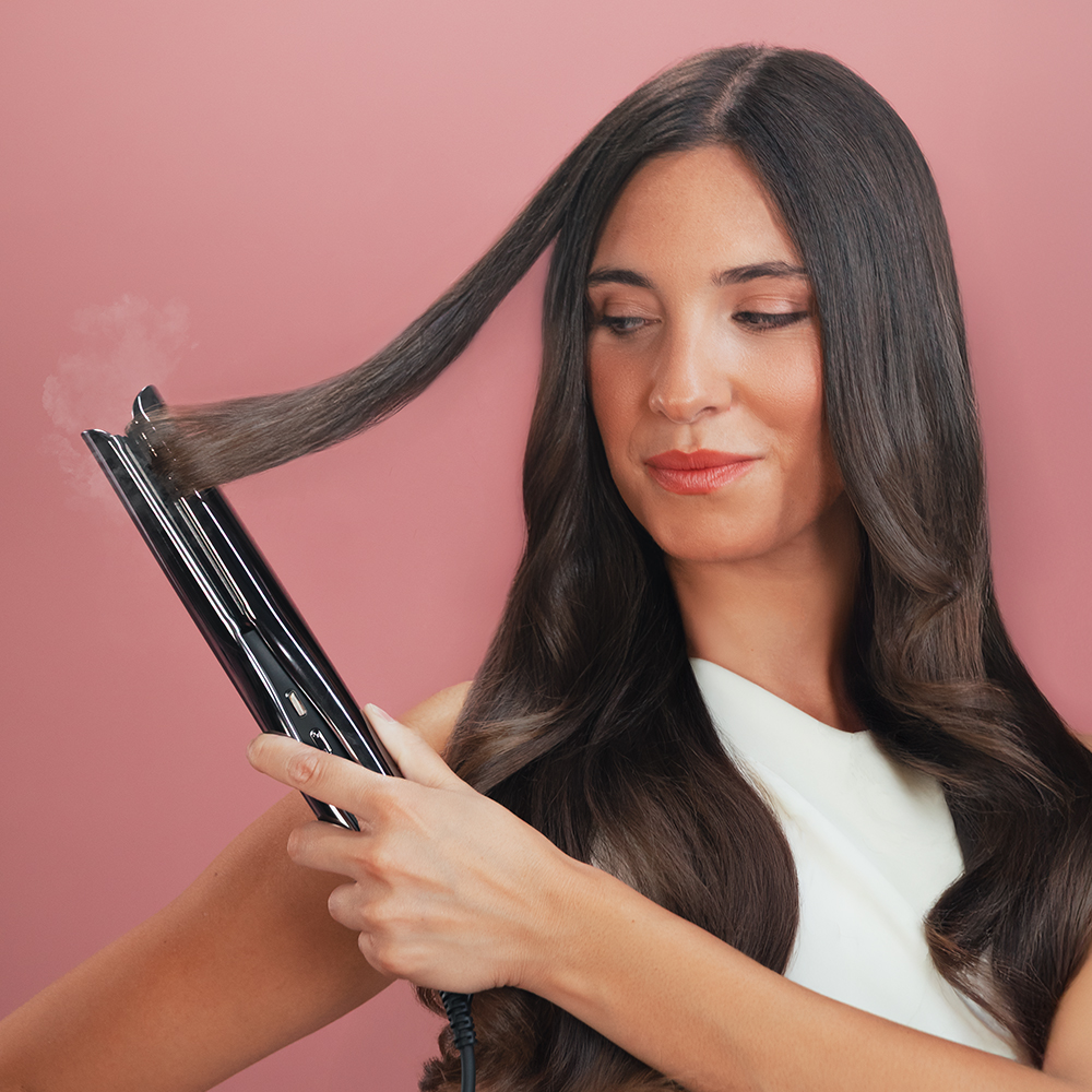 Una mujer usa una plancha de pelo negra. El fondo es rosa. Lleva una camiseta blanca.