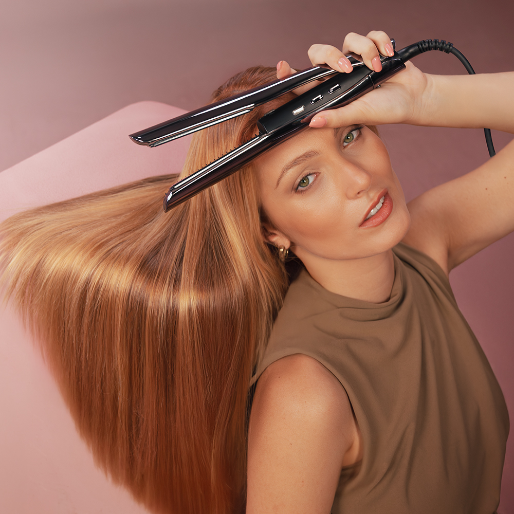 Una mujer sostiene una plancha para el pelo cerca de su cabeza. Su largo cabello rubio y liso cae. El fondo es rosa.