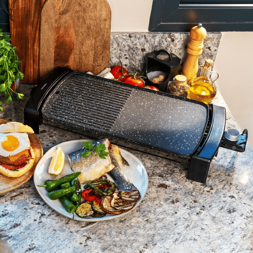 Parrilla eléctrica con pescado cocido, verduras y hamburguesa. Varios ingredientes y utensilios rodean la parrilla en una encimera de cocina.
