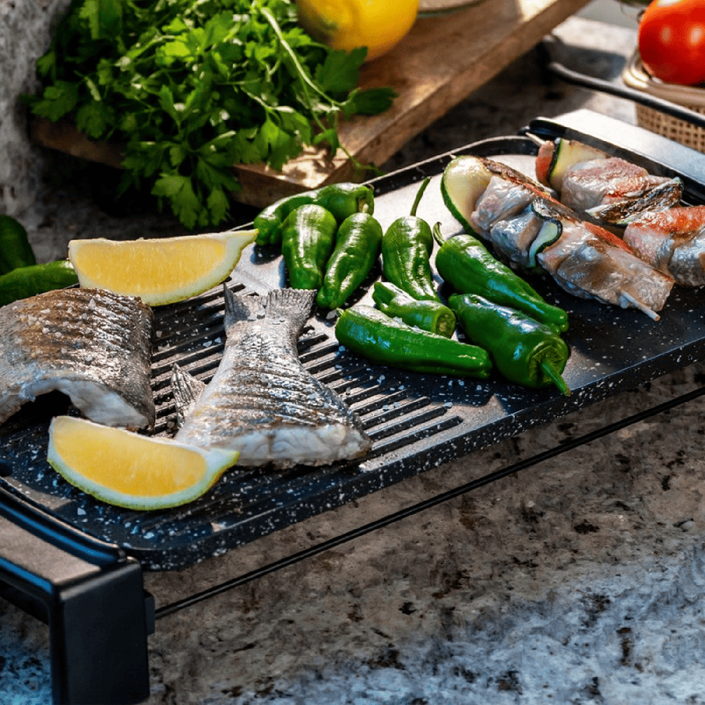 Pescado a la parrilla con pimientos verdes, rodajas de limón y brochetas en una parrilla. Perejil y un tomate al fondo.