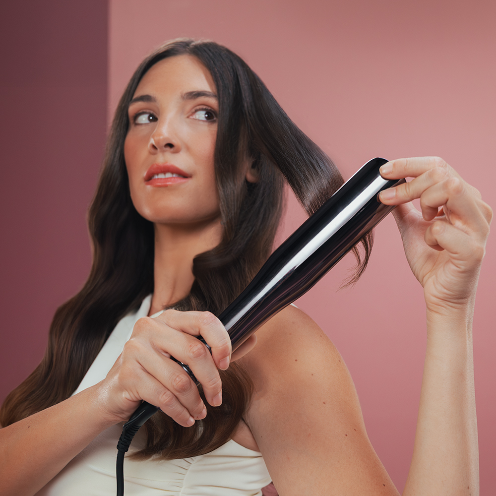 Una mujer usa una plancha de pelo negra en su largo cabello oscuro. Mira hacia un lado sobre un fondo rosa.