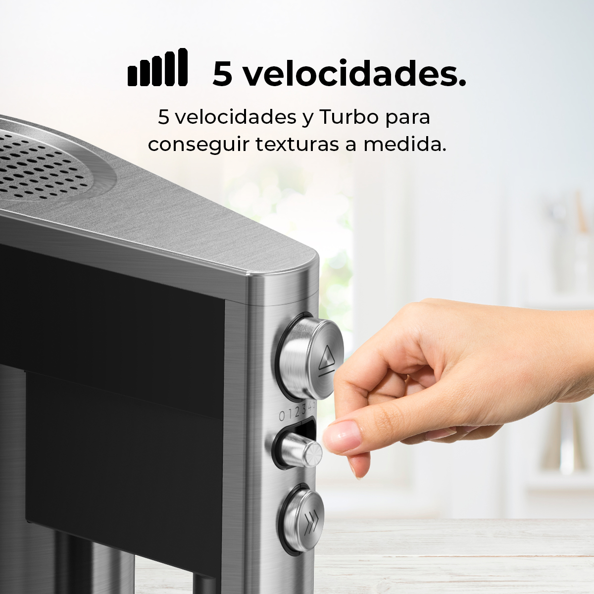 Una mano gira el dial de una batidora. La batidora es plateada con detalles en negro. El texto dice '5 velocidades'.