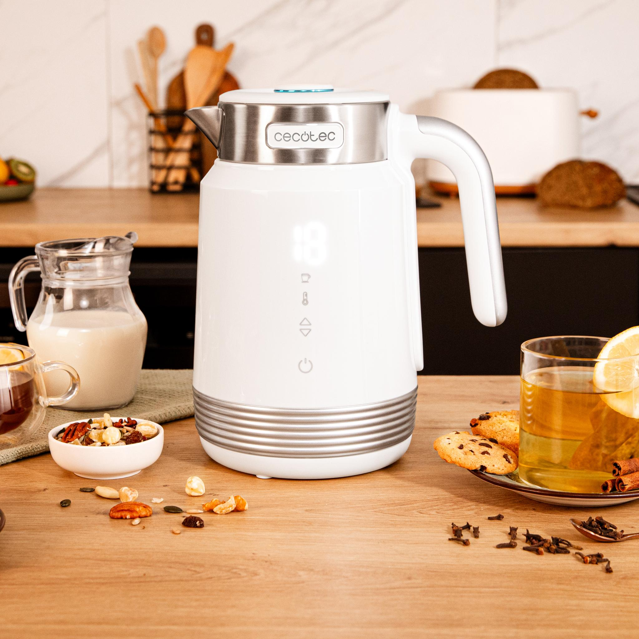 Una tetera blanca sobre mesa de madera con té, galletas, frutos secos y leche. El fondo de cocina tiene utensilios.