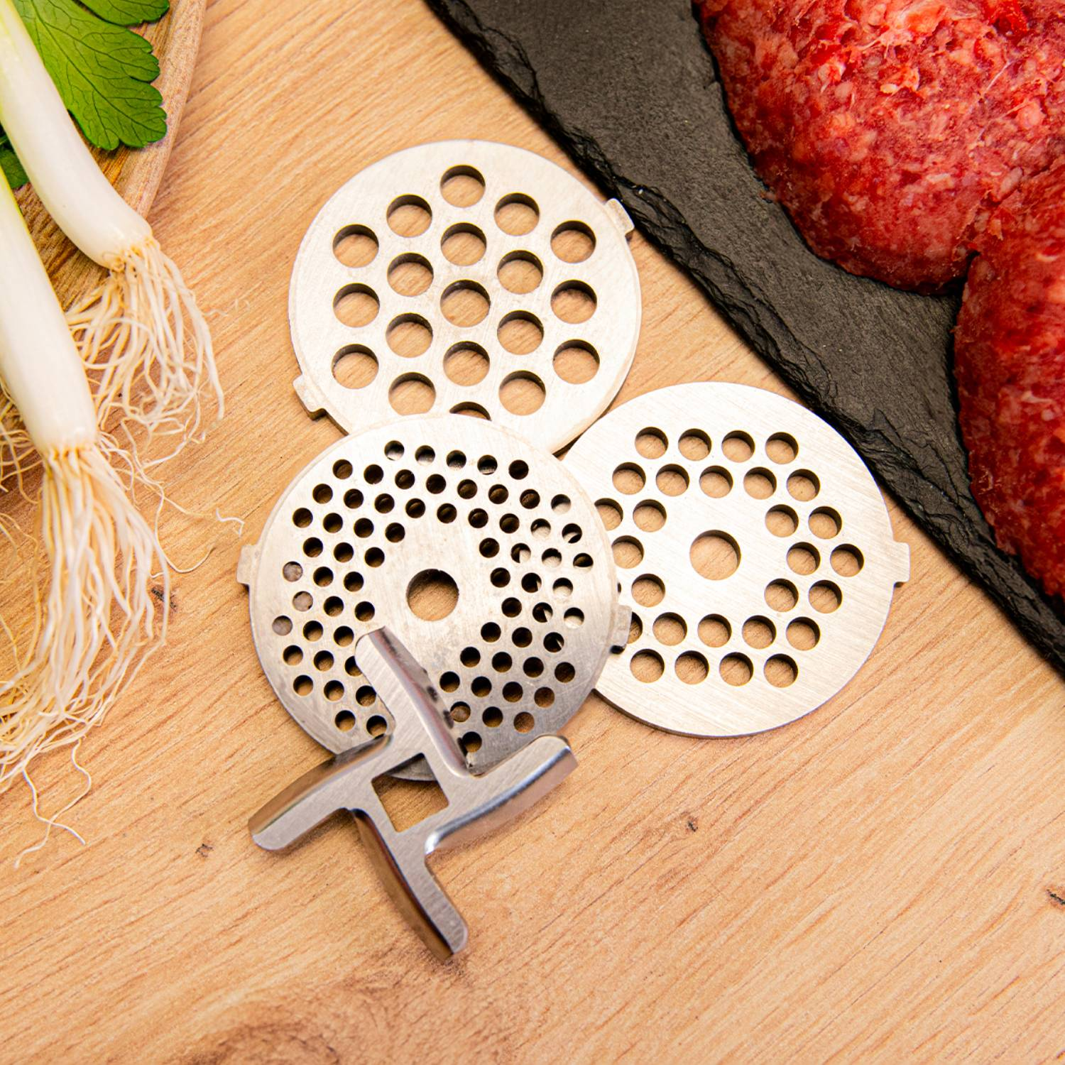 Piezas de picadora de carne con carne cruda y cebolletas sobre madera. Platos de metal y cuchilla visibles.