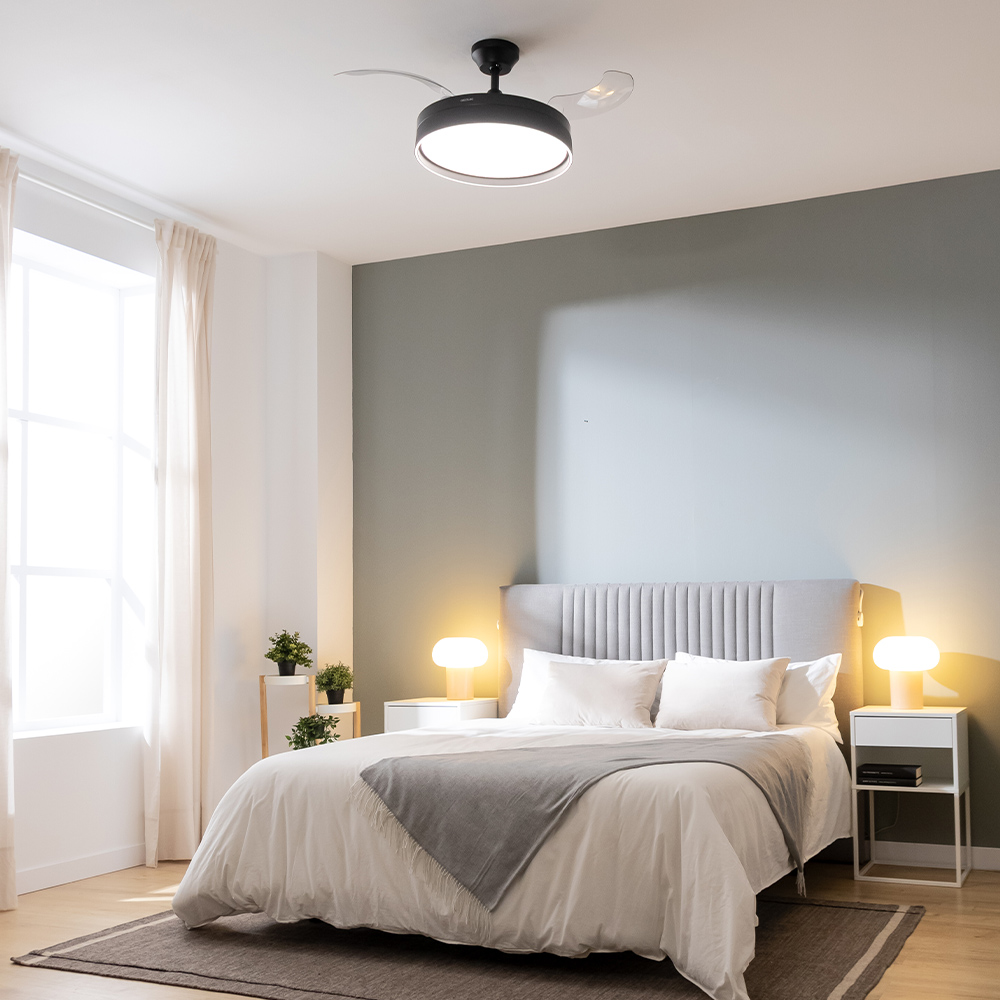 Dormitorio con cama, lámparas y ventilador de techo. La cama tiene sábanas blancas y una manta gris. Una ventana está a la izquierda.