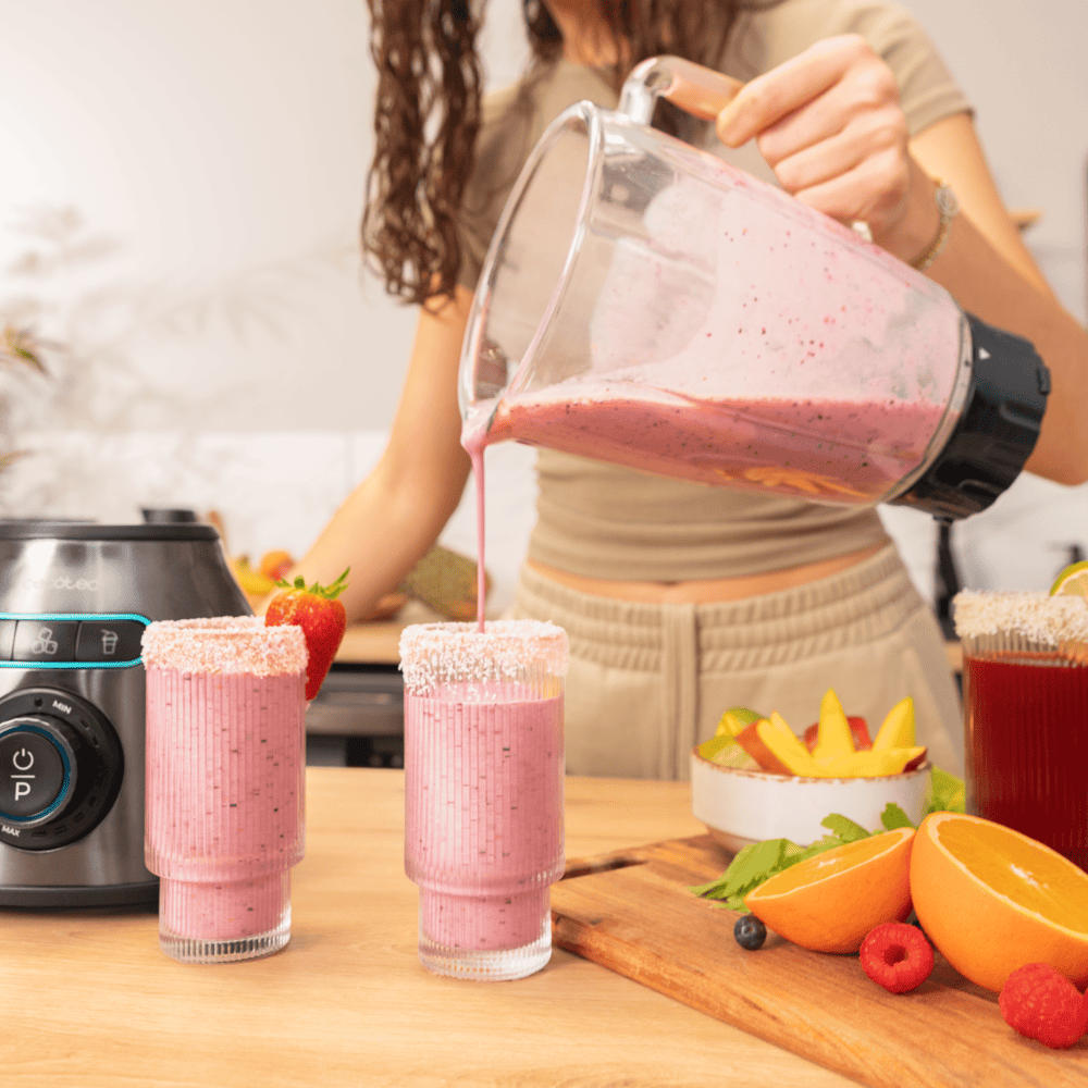 Una mujer vierte batido rosa de una licuadora en vasos. Frutas y una licuadora sobre una mesa de madera.
