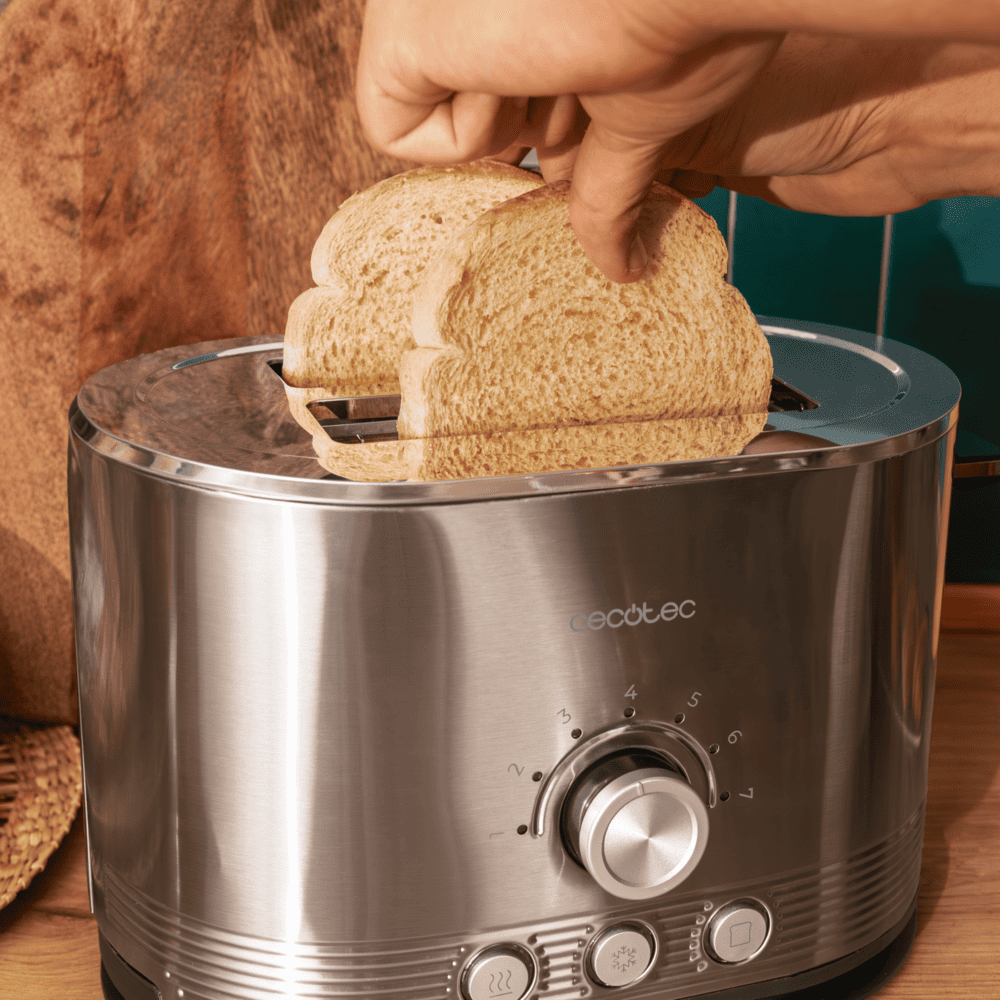 Una mano coloca dos rebanadas de pan en una tostadora cromada. Una tabla de cortar de madera y una pared verde están al fondo.