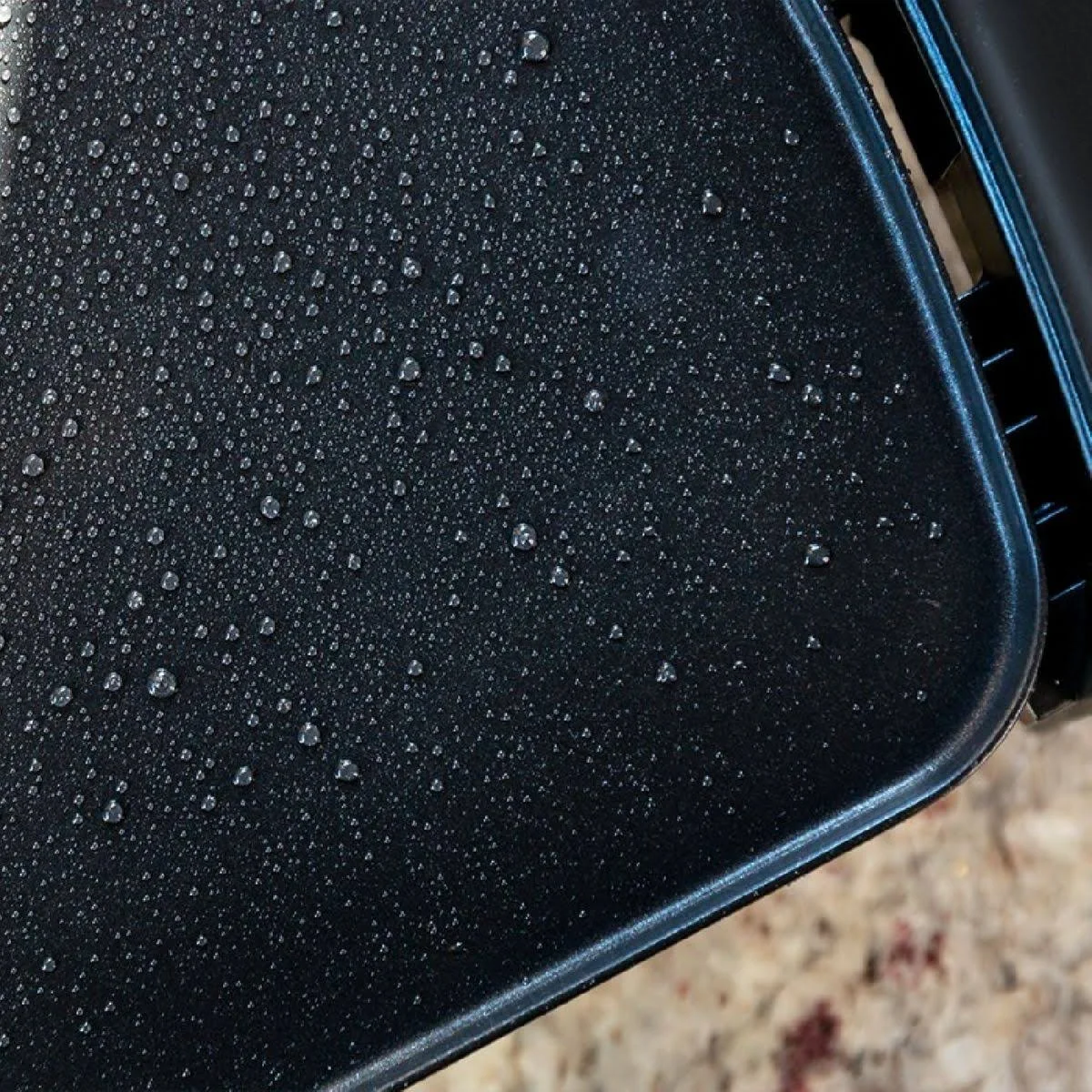 Primer plano de una plancha negra y húmeda con muchas gotas de agua. El fondo es una encimera beige moteada.