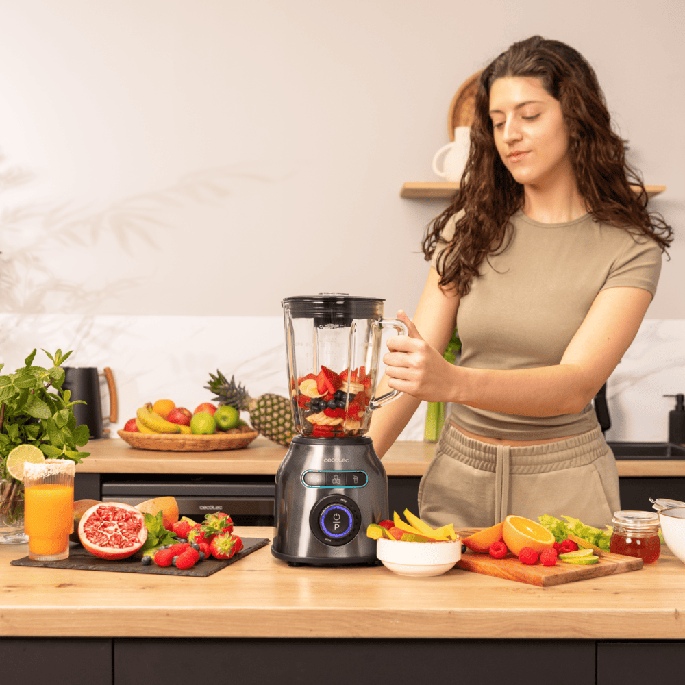 Mujer haciendo un batido con una licuadora. Se ven frutas como fresas, arándanos y plátanos. Un vaso de jugo también está en la escena.