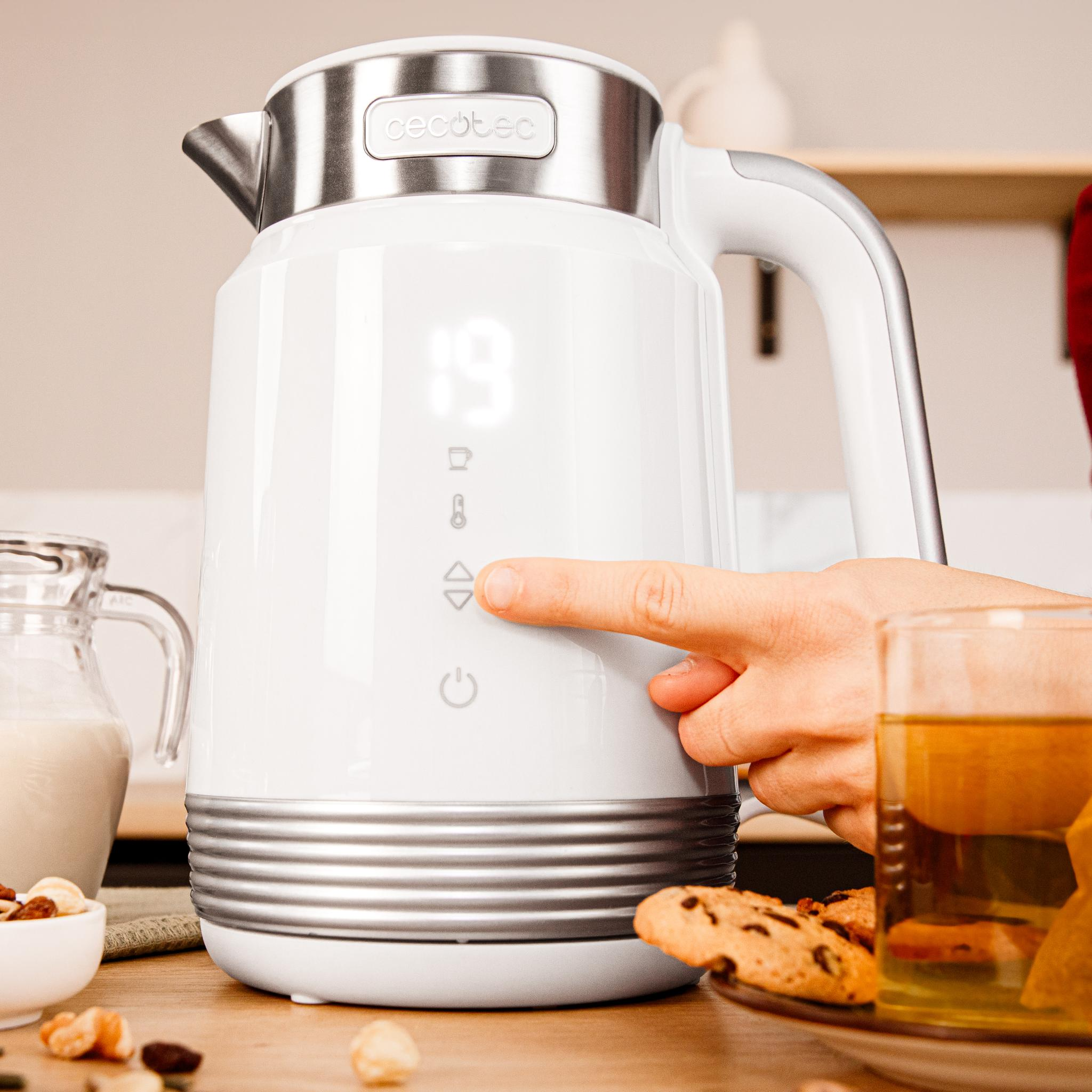 Una mano presiona un botón en una tetera eléctrica blanca. Un vaso de té, galletas y nueces están en la mesa.