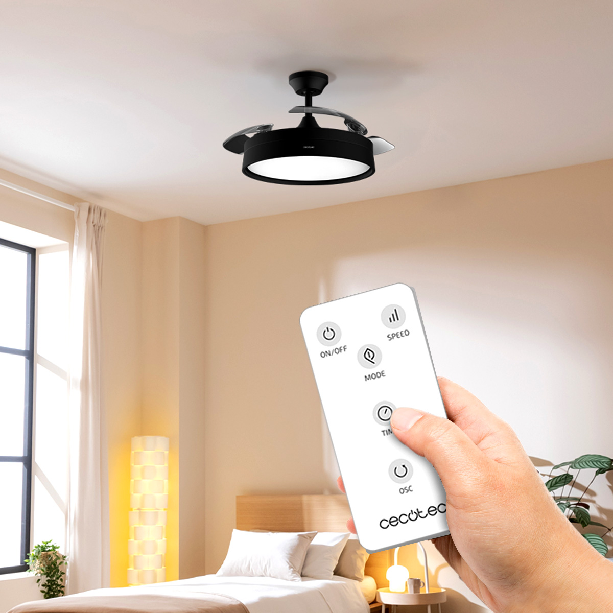 Una mano sostiene un mando a distancia que apunta a un ventilador de techo con luz en un dormitorio. La habitación tiene una cama, una lámpara y una ventana.