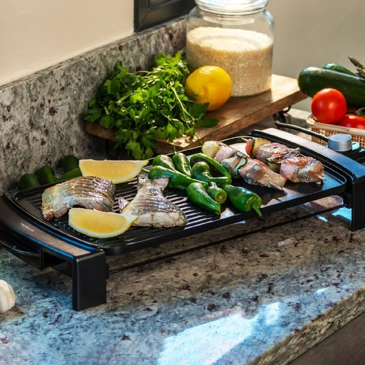 Una escena de cocina con pescado a la parrilla, verduras y limón en una parrilla negra. También hay otros ingredientes como arroz, tomates y perejil.