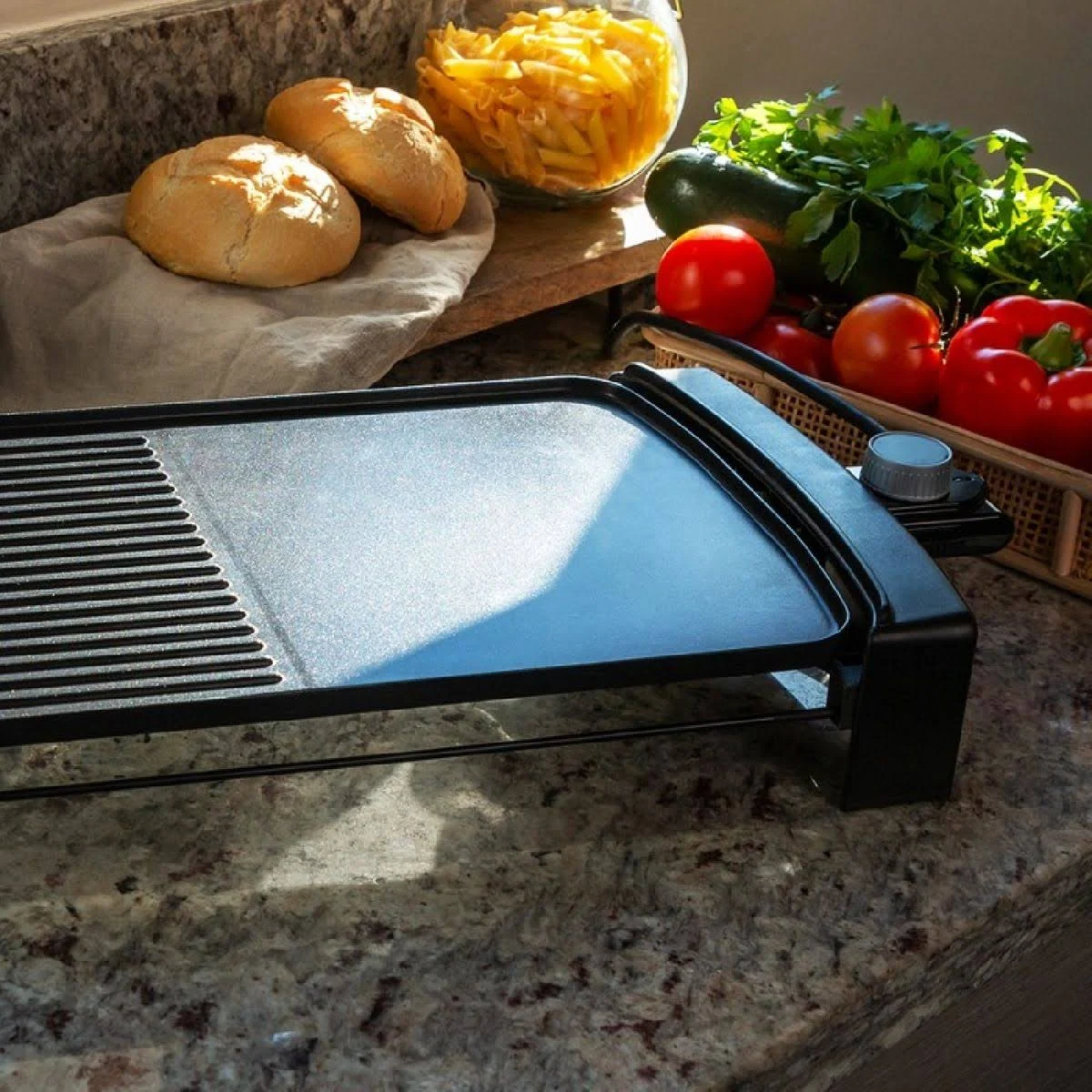 Parrilla eléctrica en mostrador con pan, pasta, tomates, pimientos y calabacín. La luz solar ilumina la escena.