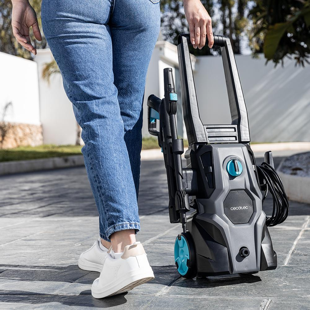 Una persona camina, llevando una hidrolavadora. La lavadora es negra y gris con detalles turquesa. La persona lleva vaqueros y zapatillas blancas.