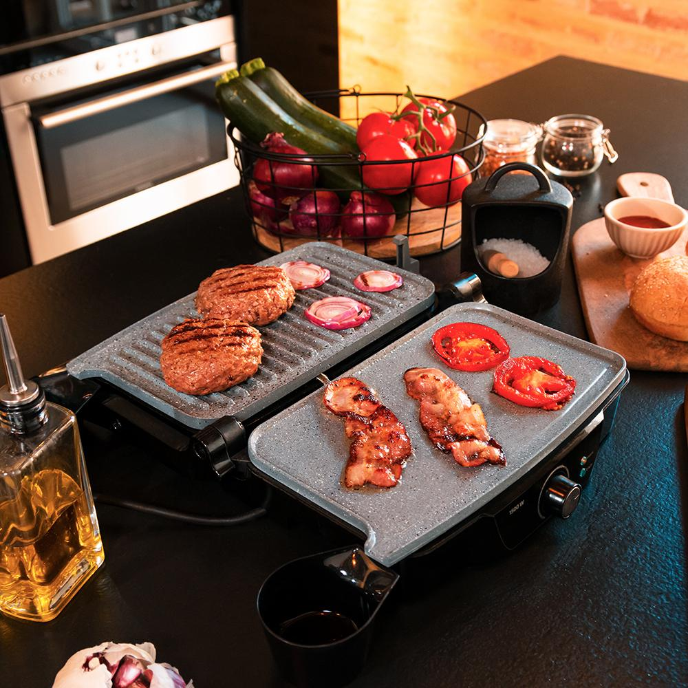 Escena de cocina con dos parrillas cocinando hamburguesas, tocino y tomates. Verduras en una cesta, aceite y sal cerca.