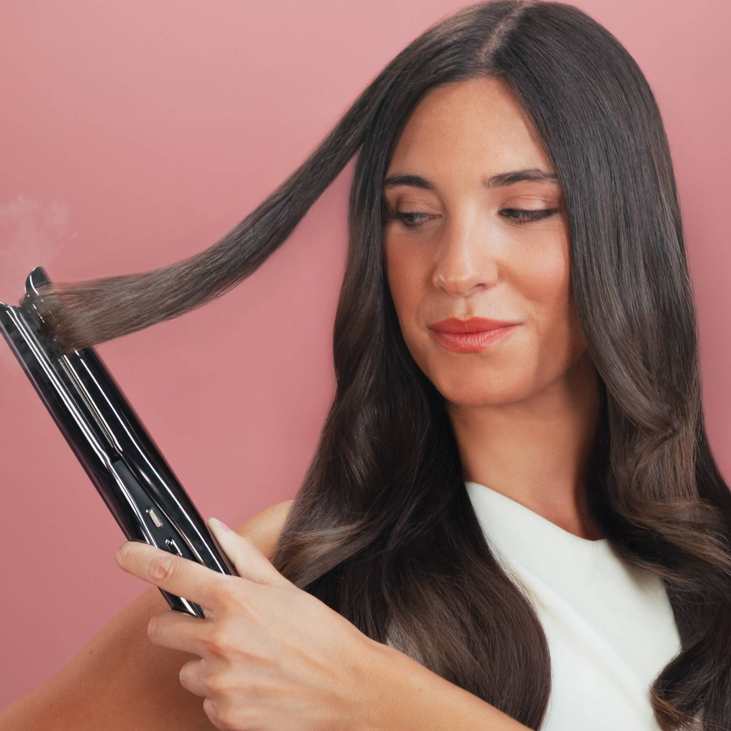 Una mujer usa una plancha de pelo negra en su largo cabello oscuro sobre un fondo rosa.