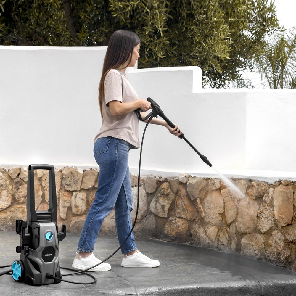 Una mujer en vaqueros y camiseta utiliza una hidrolavadora en un patio. Máquina a la izquierda. Muro de piedra y pared blanca en el fondo.