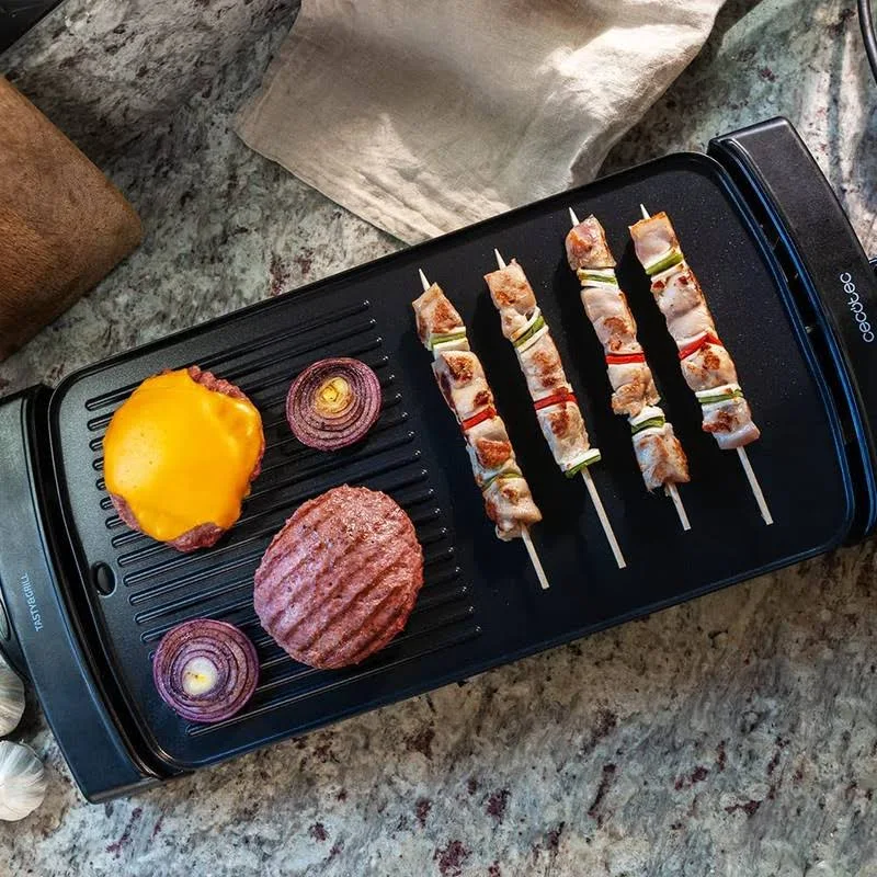 Una parrilla negra con brochetas, una hamburguesa con queso y cebollas rojas. Un paño beige y una tabla de madera están en el fondo.