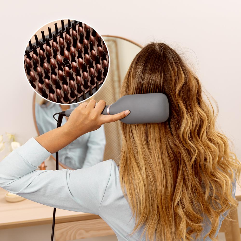 Una mujer se cepilla su cabello rubio y ondulado con un cepillo térmico gris. Un recuadro circular muestra el detalle del cepillo. Un espejo al fondo.