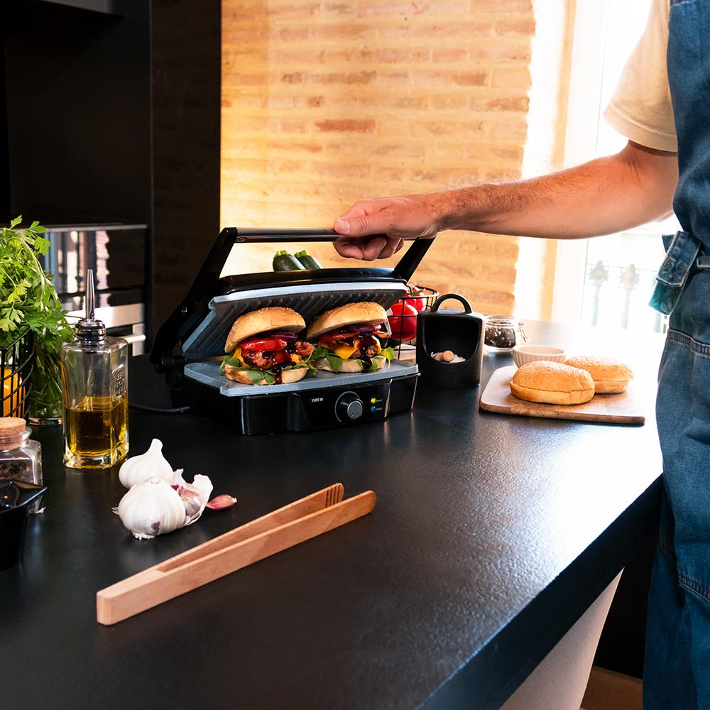 Una persona está asando hamburguesas en una parrilla negra. Ajo, pinzas, aceite y panecillos en la encimera. Pared de ladrillos al fondo.