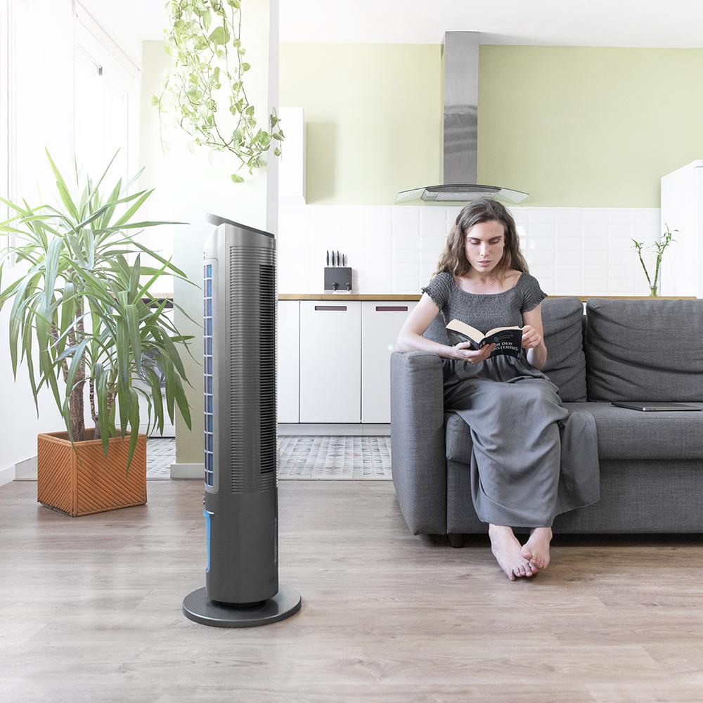 Una mujer lee en un sofá. Un ventilador y una planta grande están en primer plano. La cocina está al fondo.