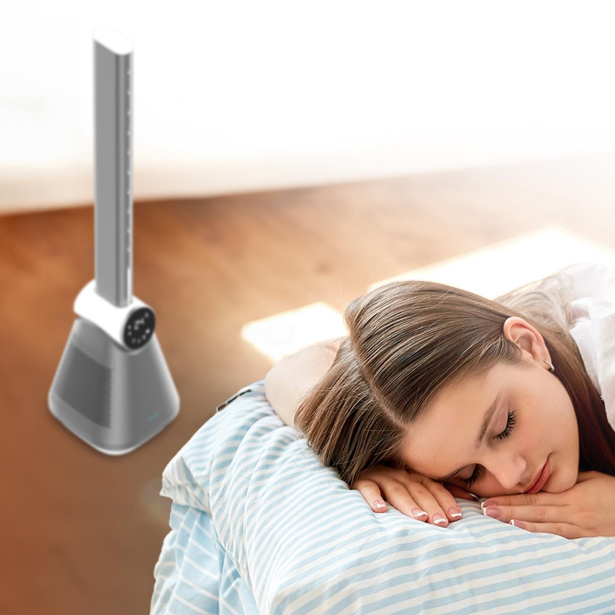 Una mujer duerme en una cama con una manta a rayas azules, cerca de un ventilador de torre gris. La habitación tiene pisos de madera.