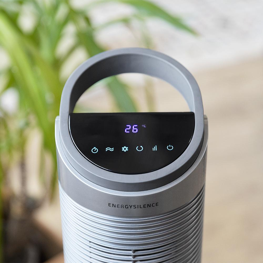 Un purificador de aire gris muestra 26 grados. Tiene un asa y un panel de control con iconos. Planta verde al fondo.