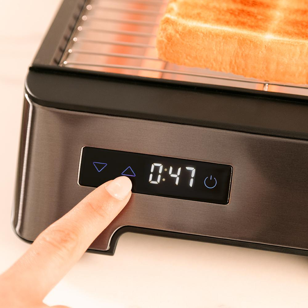 Una mano toca la pantalla digital de una tostadora mostrando 0:47. Una rebanada de pan tostado está en la tostadora.