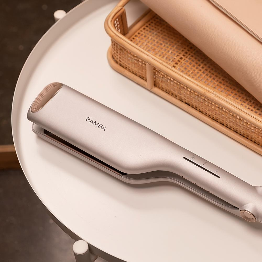 Una plancha de pelo plateada con la etiqueta 'BAMBA' sobre una mesa blanca. Una cesta de mimbre y un embrague beige al fondo.