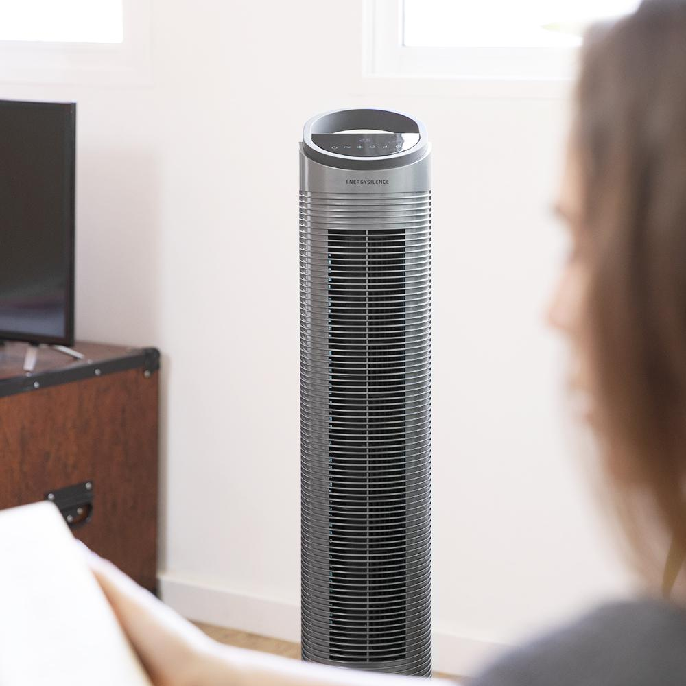 Una mujer mira un ventilador de torre gris. El ventilador está frente a una pared blanca y una ventana. También se ve un televisor y un mueble de madera.