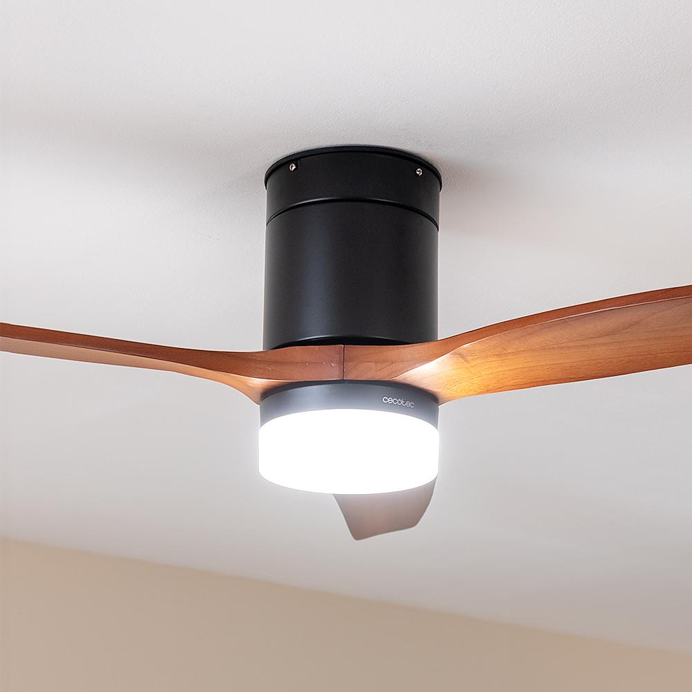 Ventilador de techo con aspas de madera. Tiene un motor negro y una lámpara. El fondo es un techo blanco y una pared beige.