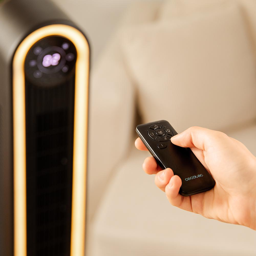Una mano sostiene un mando a distancia negro, operando un ventilador negro y dorado. Un sofá está en el fondo borroso. El mando tiene el logo Cecotec.