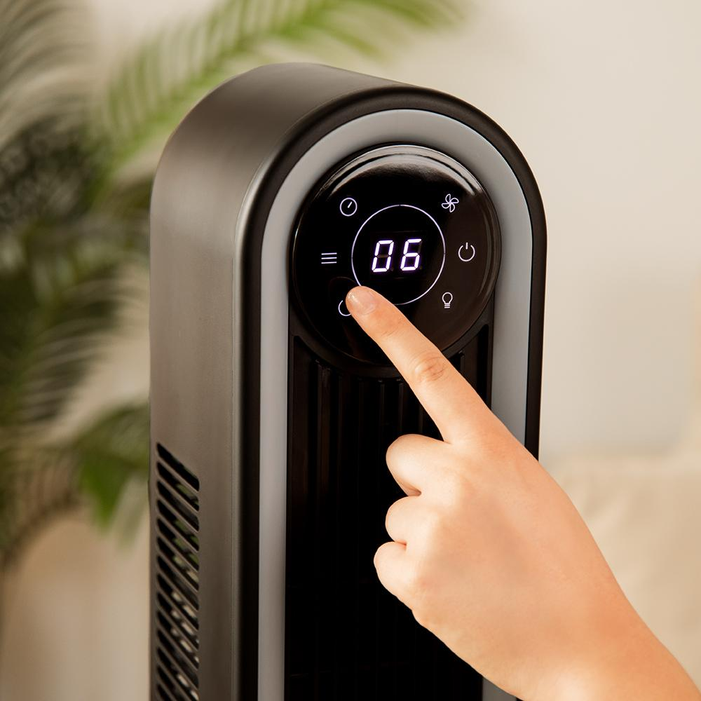 Una mano toca el panel de control del ventilador mostrando '06'. El ventilador negro tiene un borde plateado. El fondo es una planta.