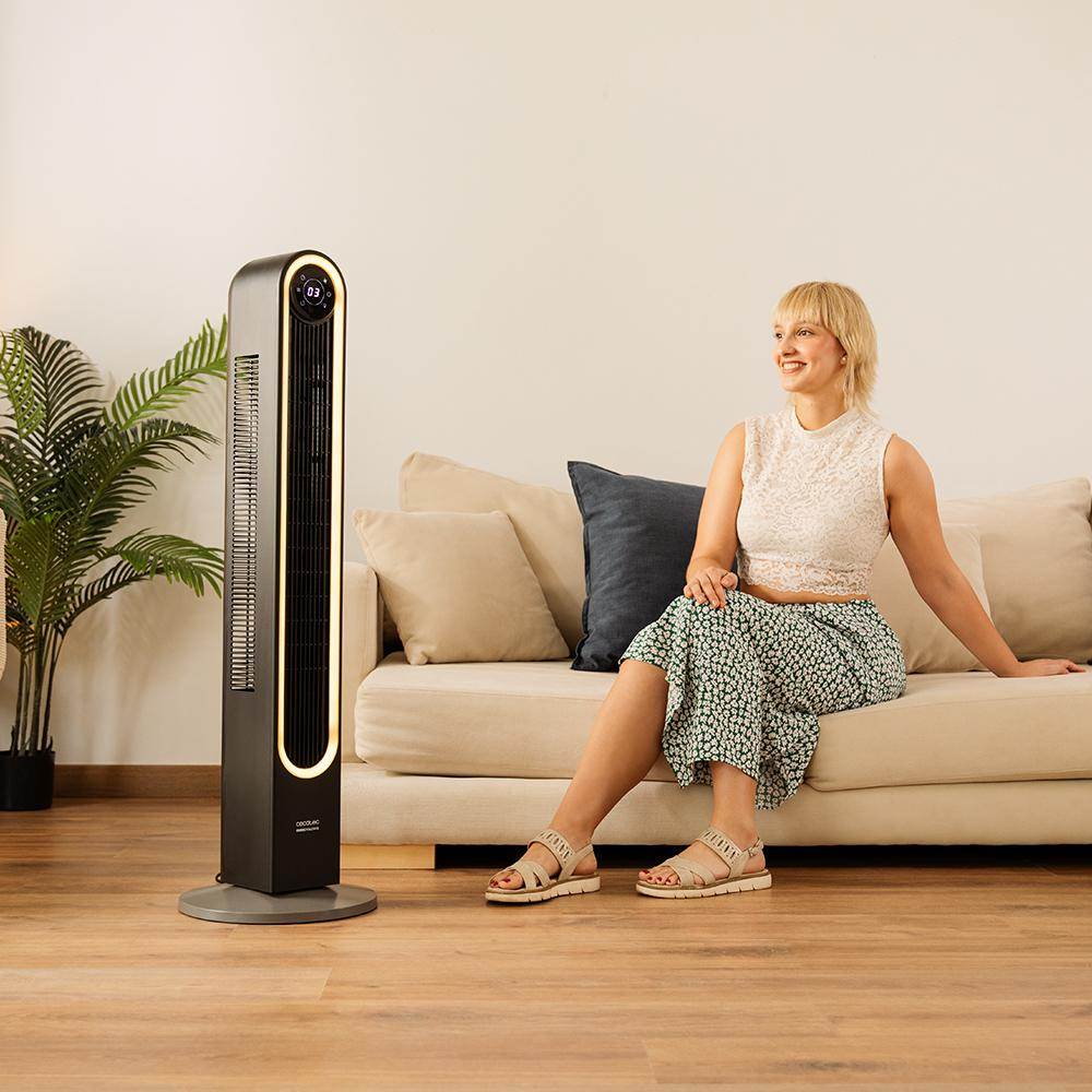 Una mujer sentada en un sofá junto a un ventilador negro. Lleva un top blanco y una falda floral. Hay una planta a la izquierda.