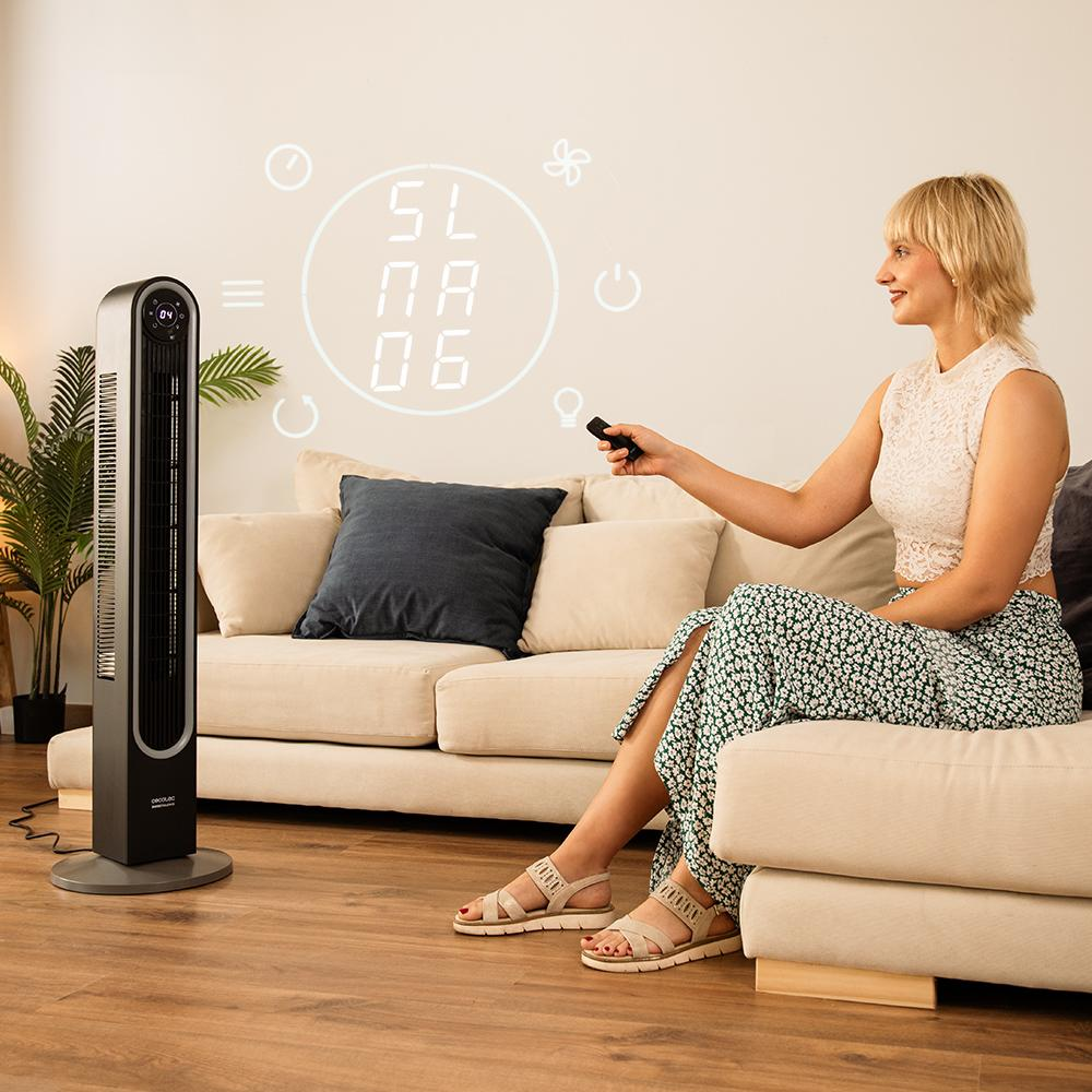 Mujer con mando a distancia controlando un ventilador. Está sentada en un sofá. Números proyectados en la pared. Planta y ventilador visibles.