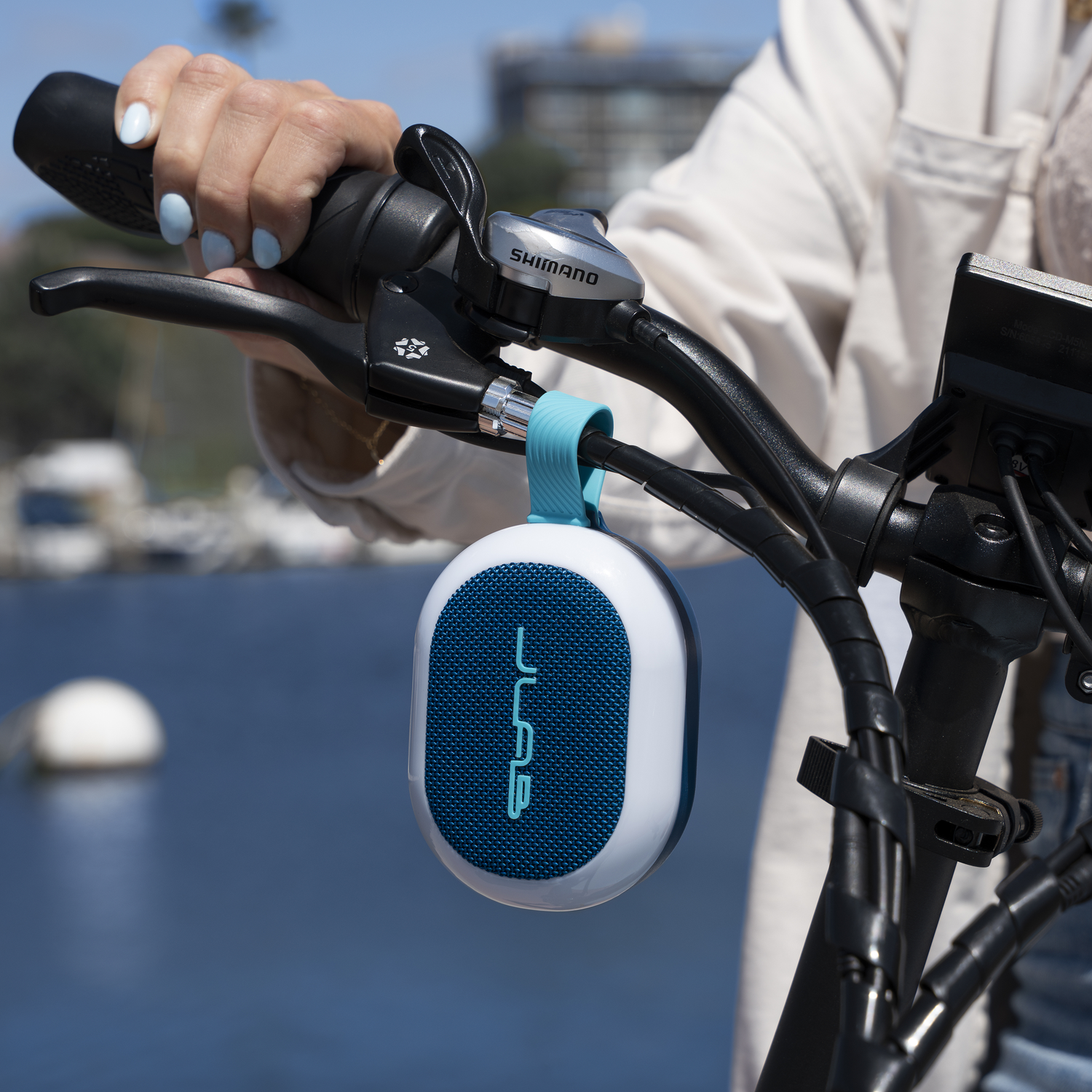 A person's hand on a bike handle, holding a speaker. Blue water and a blurry background of boats and buildings are visible.
