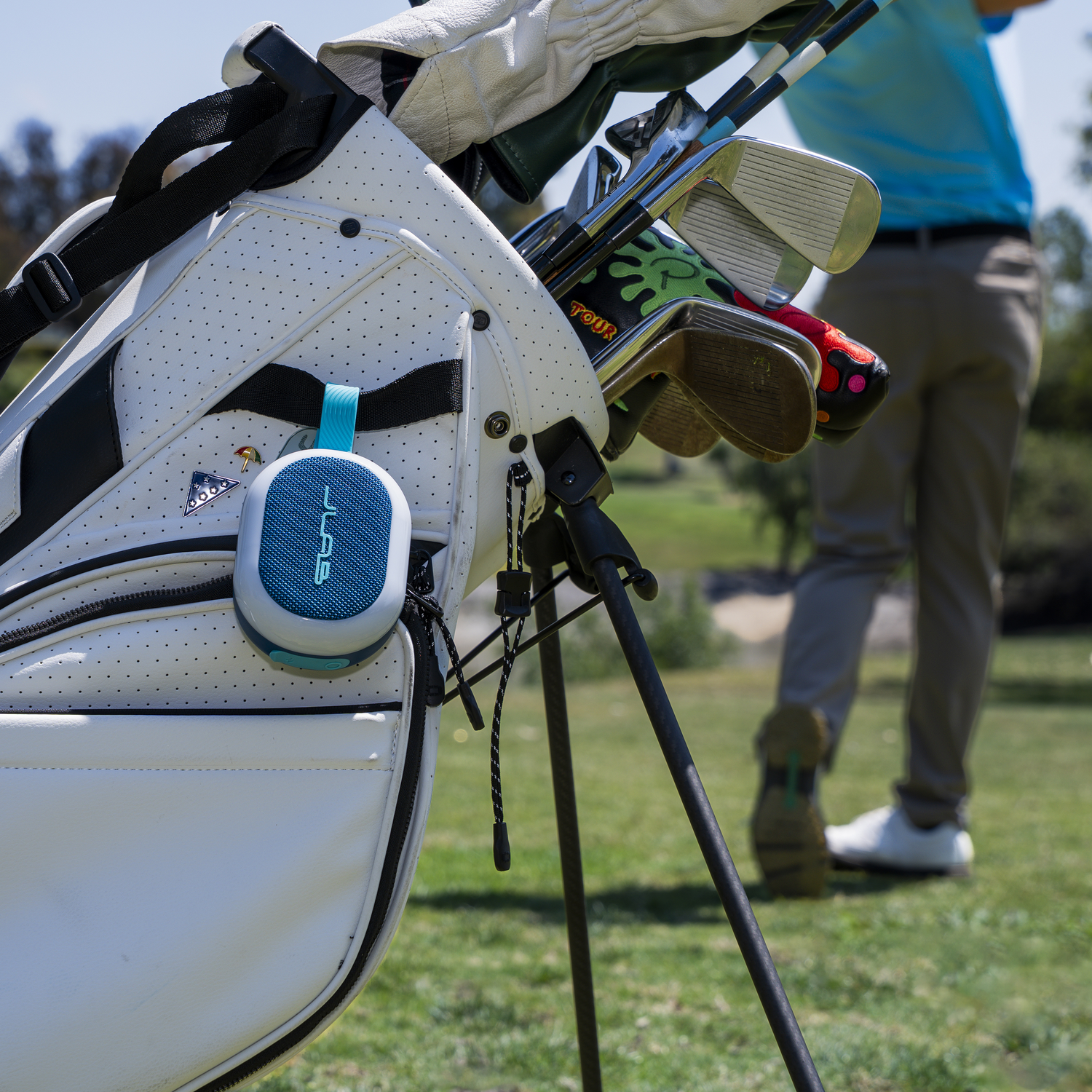 A golf bag with clubs and a speaker attached. A golfer stands in the background on a green golf course.