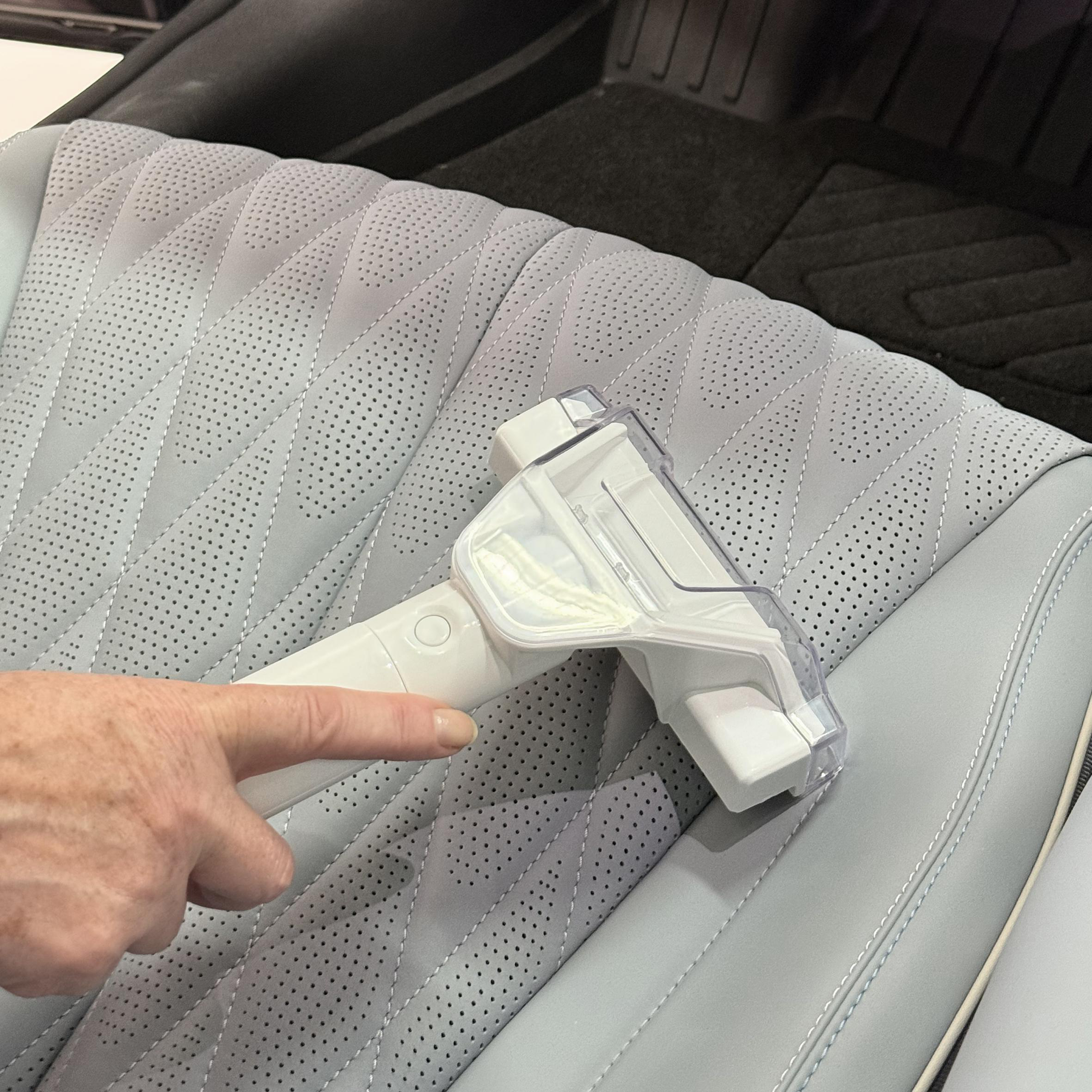 Eine Hand zeigt auf einen weißen und transparenten Handstaubsauger auf einem grauen perforierten Autositz.