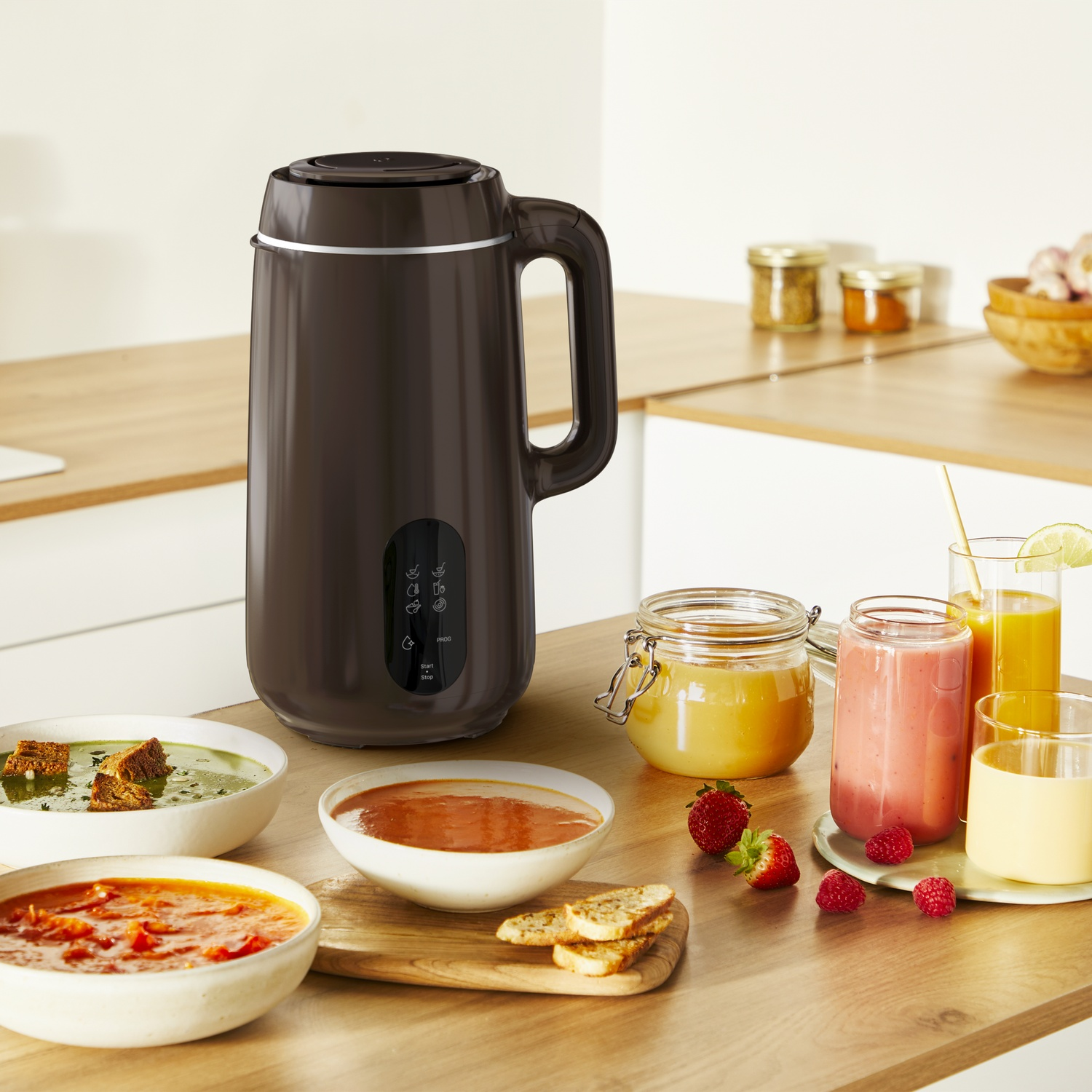 A brown soup maker with bowls of soup, smoothies, and ingredients on a wooden table. Background has jars and a wooden countertop.