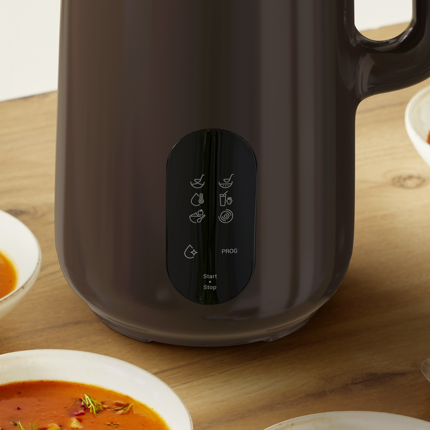 A brown soup maker on a wooden table. Bowls of soup surround it. The maker has a control panel with icons and buttons.