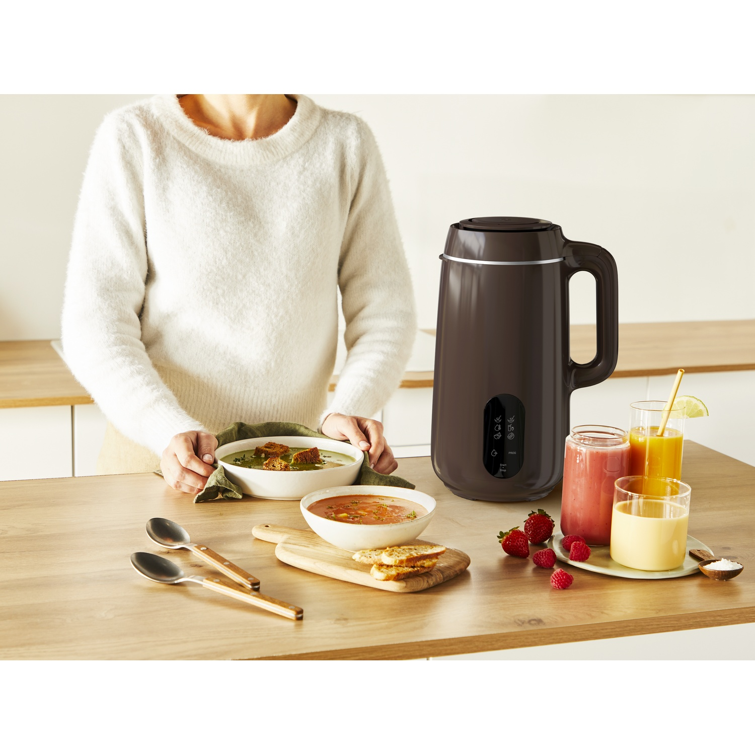 A woman stands behind a table with soup bowls, juice glasses, and a dark brown blender. Fresh fruits and spoons are present.