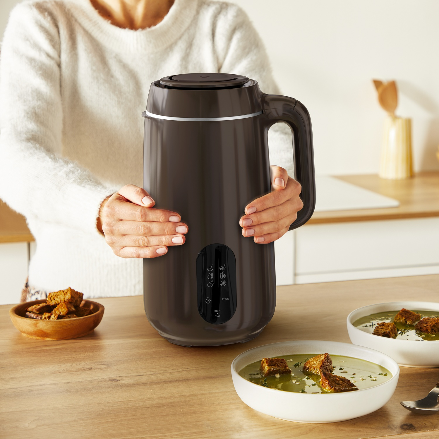 A person holds a black soup maker. Bowls of soup with croutons and a bowl of croutons sit on a wooden counter.