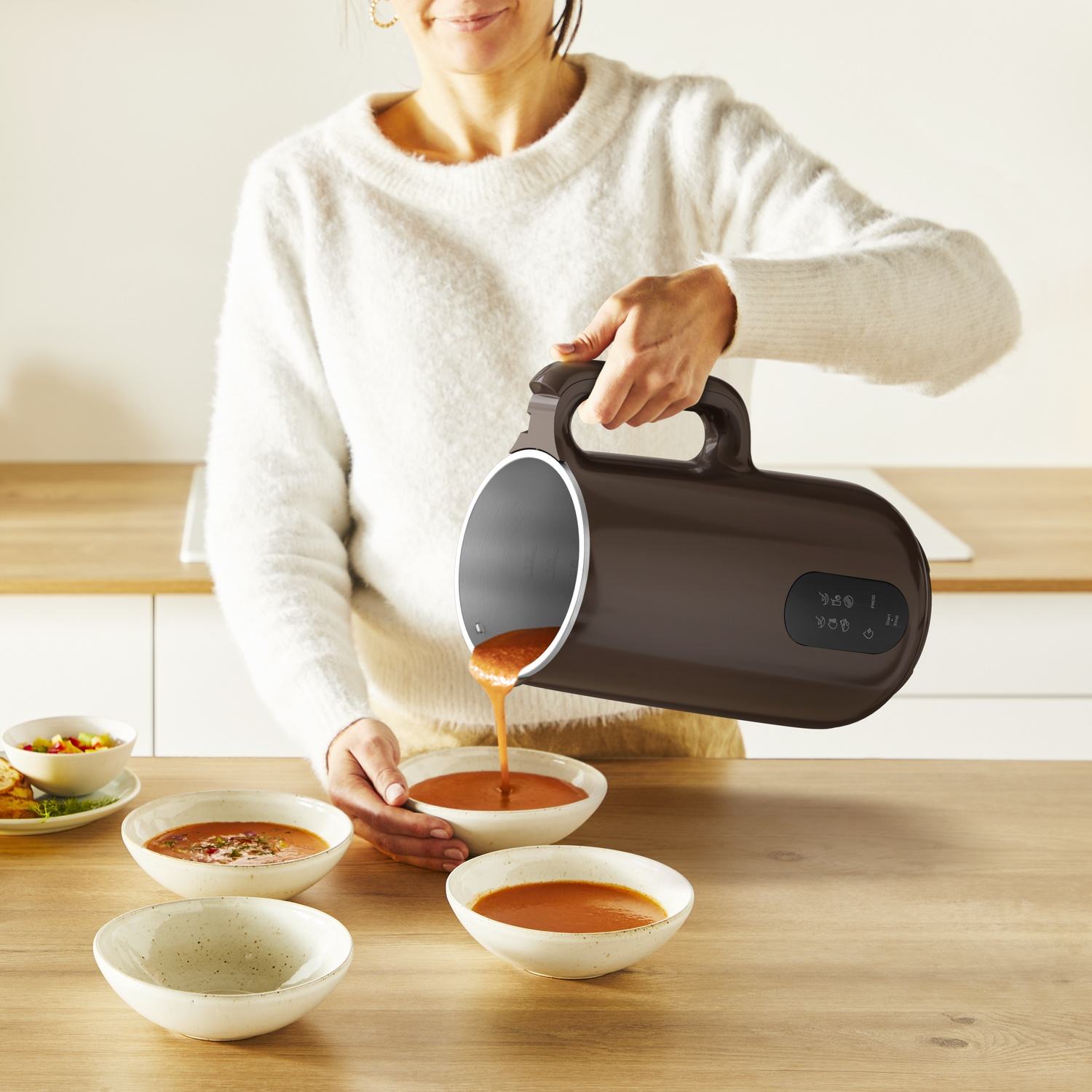 A woman pours soup from a brown pitcher into white bowls. Other bowls with soup are on a wooden table.
